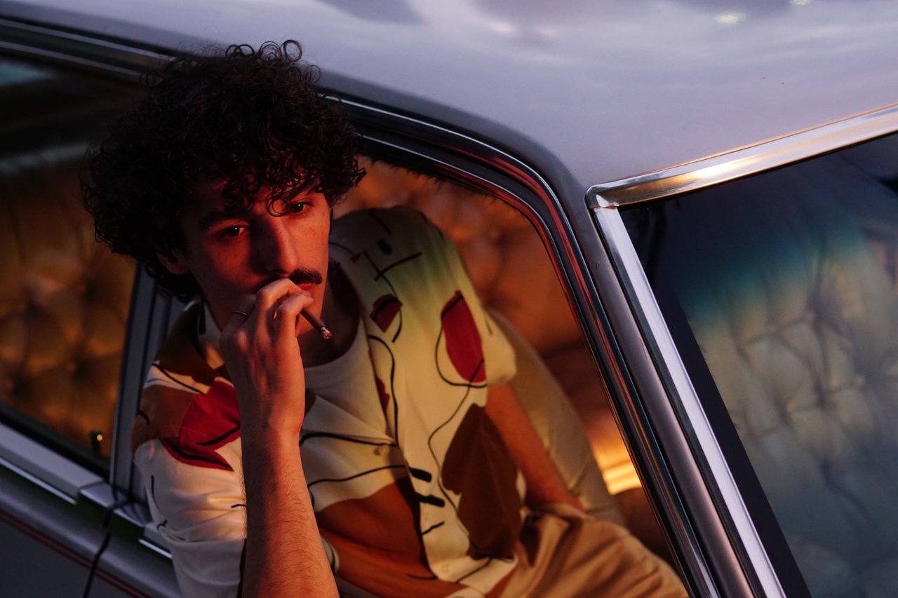 Man with curly hair and mustache smoking in a vintage car during twilight, creating a moody ambiance.