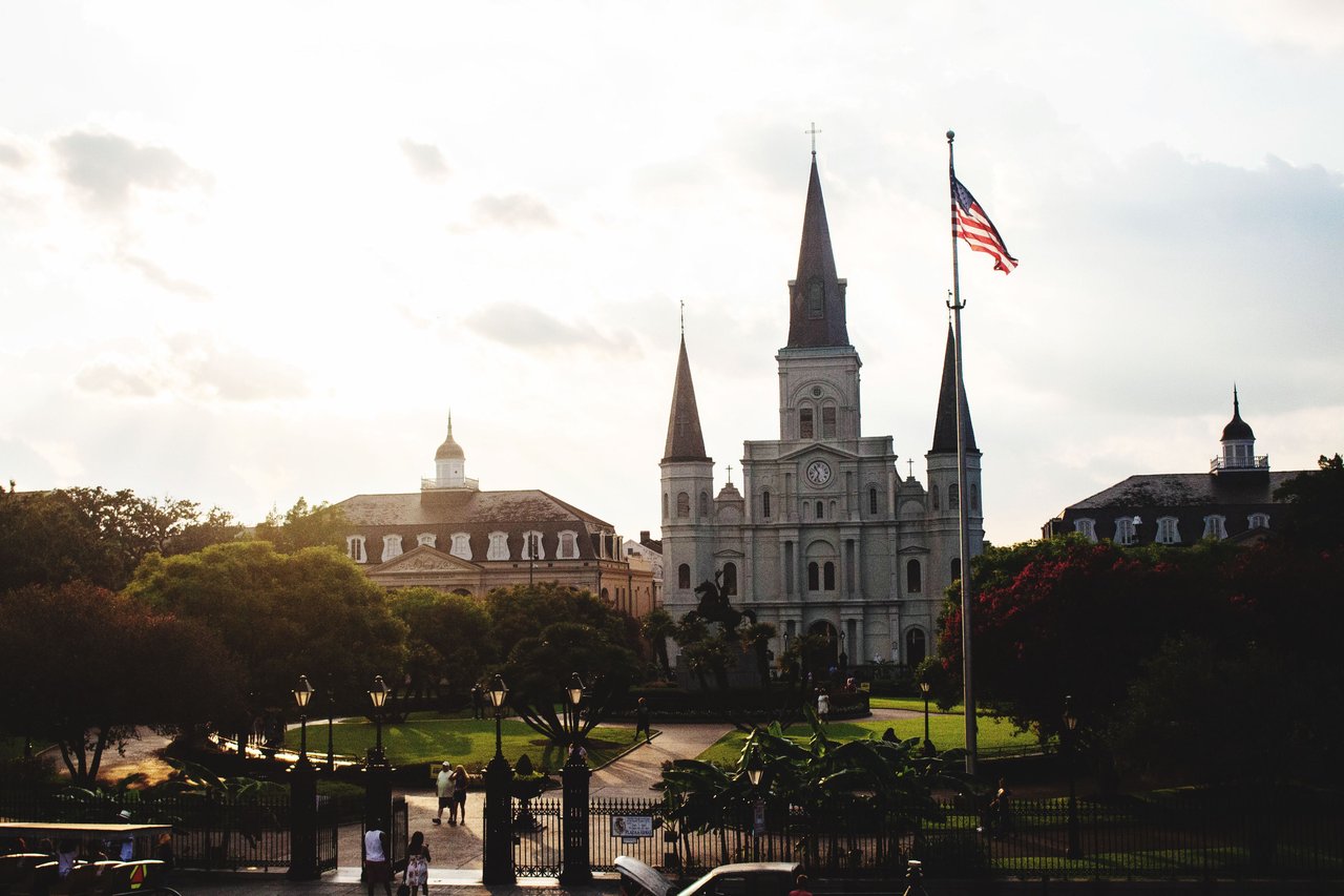 St. Louis Cathedral in New Orleans with an American flag, trees, and a garden at dusk. Iconic religious landmark.