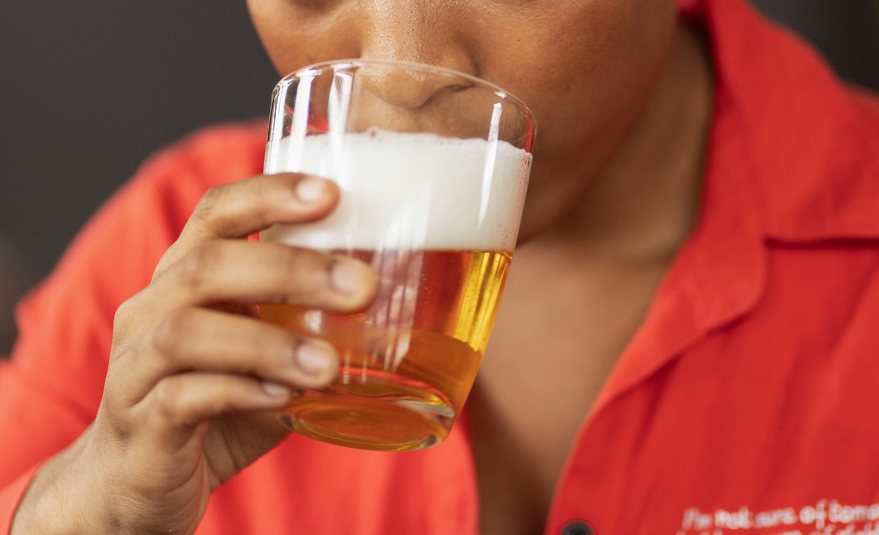 A person in a red shirt taking a sip of beer from a glass.