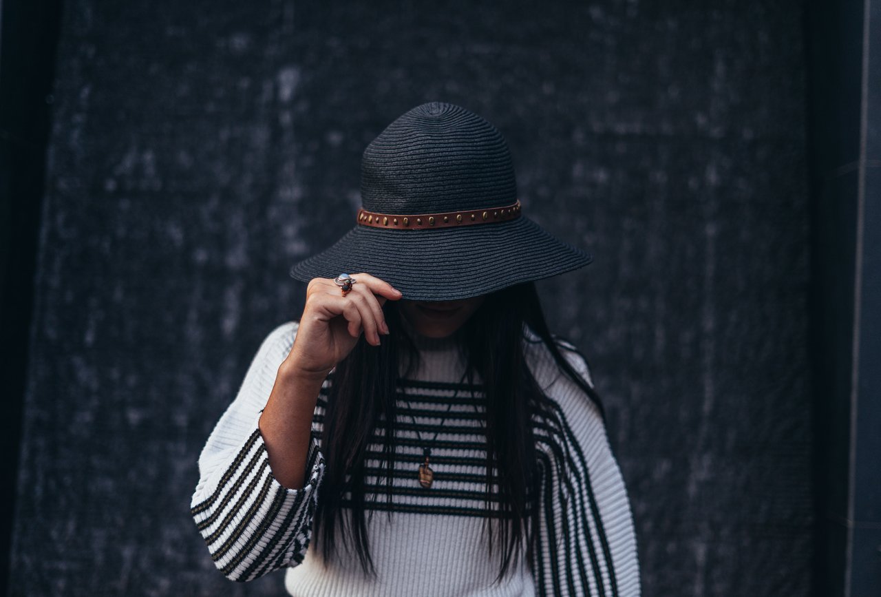 A fashionable woman wearing a stylish black hat and striped sweater stands with her face partially hidden.