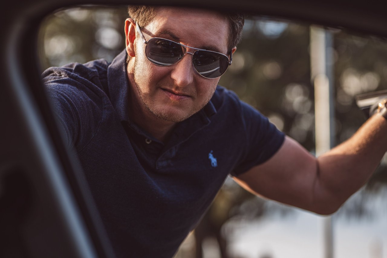 A fashionable man with aviator sunglasses leaning on a car, exuding a cool retro vibe.