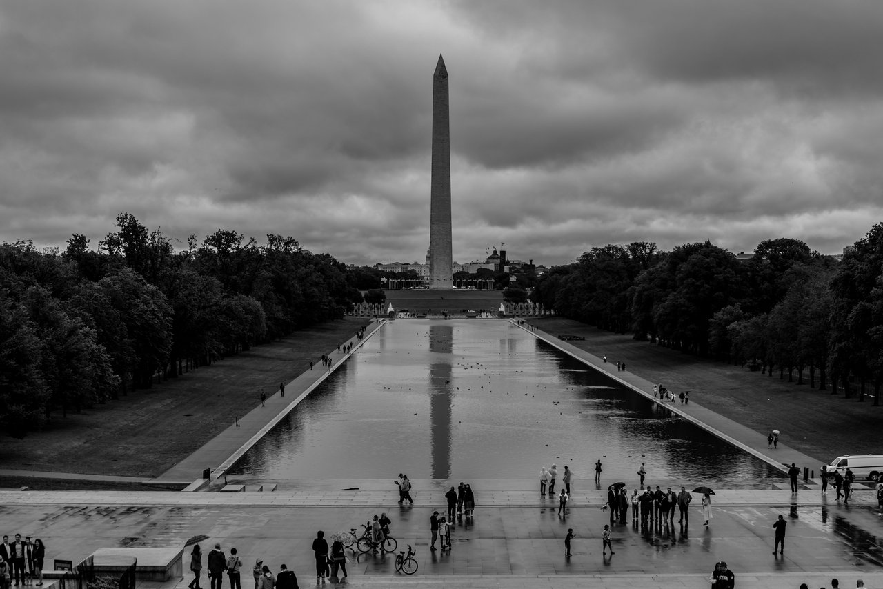 The Washington Monument stands tall reflected in the wet plaza after rain on a cloudy day.